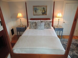 Un lit à baldaquin avec un couvre-lit blanc et de nombreux oreillers, flanqué de deux tables de chevet avec des lampes, dans une pièce dotée d’un plancher en bois et d’une porte visible au fond.