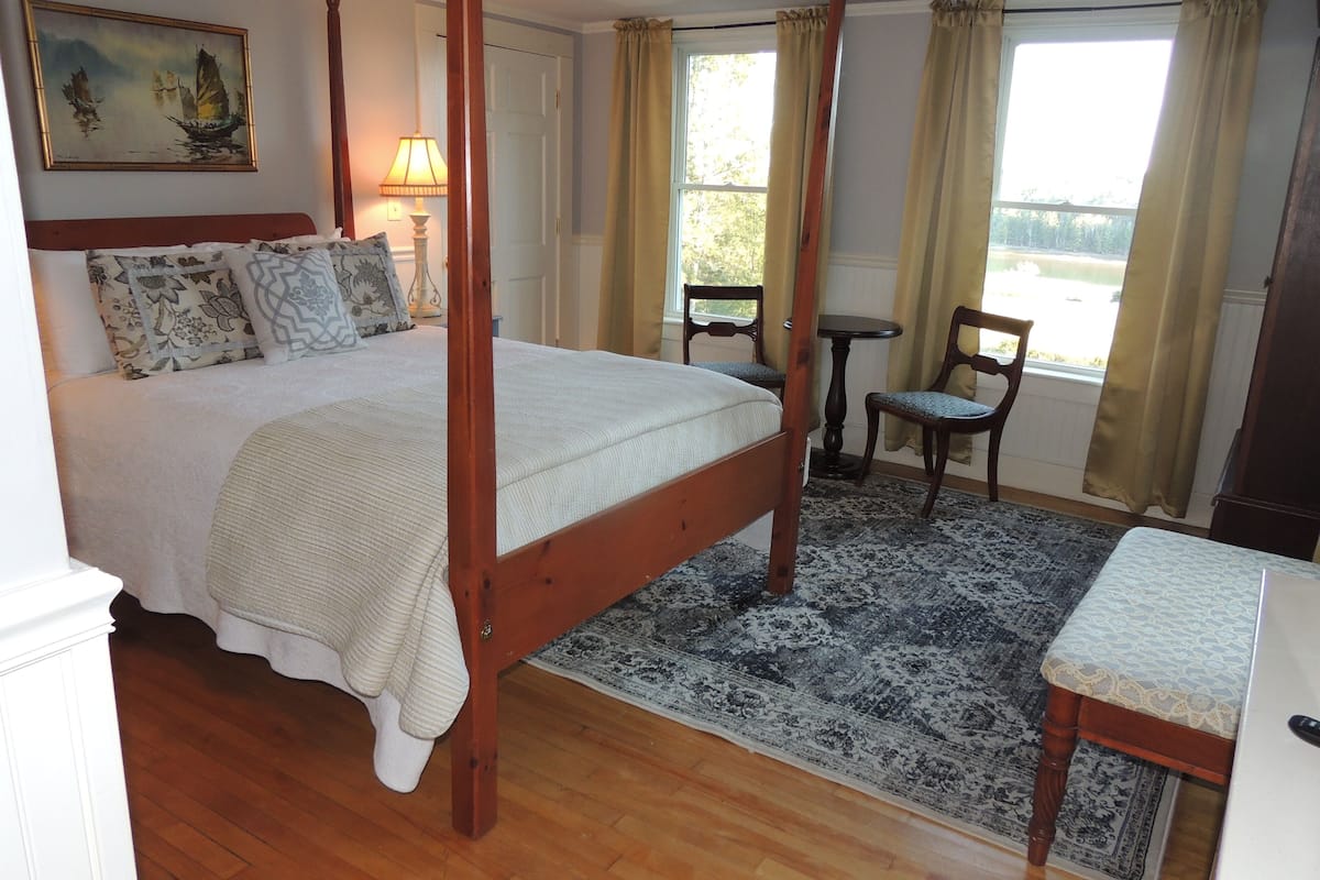 A four-poster bed with a wooden frame, a large painting on the wall, and a view of the outdoors through the window.