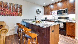 A modern kitchen with wooden cabinets, a granite countertop, a stainless steel microwave, and a bar area with stools. - Estes Park Condo Rental: Balcony w/ River Views! (Estes Park)