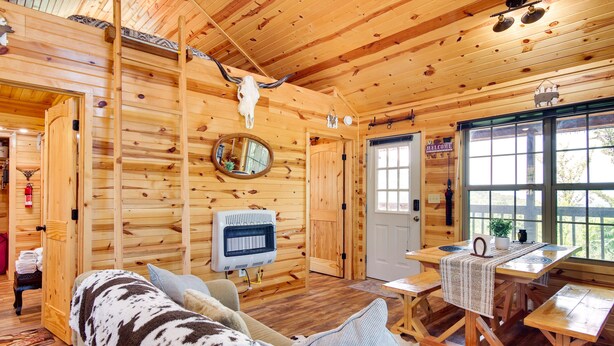 Ein Holzblockhaus-Interieur mit Holzbalkendecke, Kamin, einer Couch, einem Esstisch und einem großen Fenster.