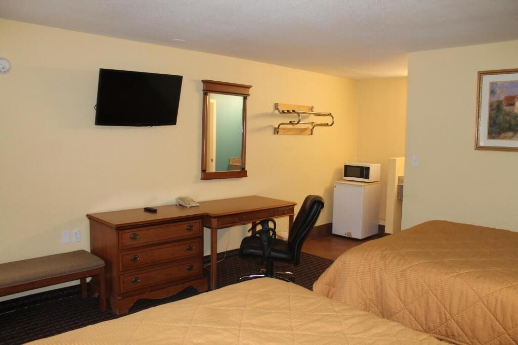 A hotel room with a bed, a desk with a chair, a television, a mirror, and a small refrigerator.