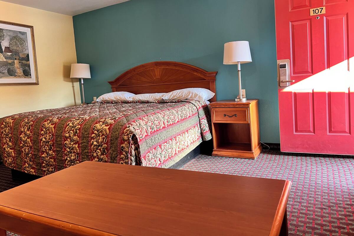 A hotel room with a bed, nightstand, lamp, and a red door with the number 107.
