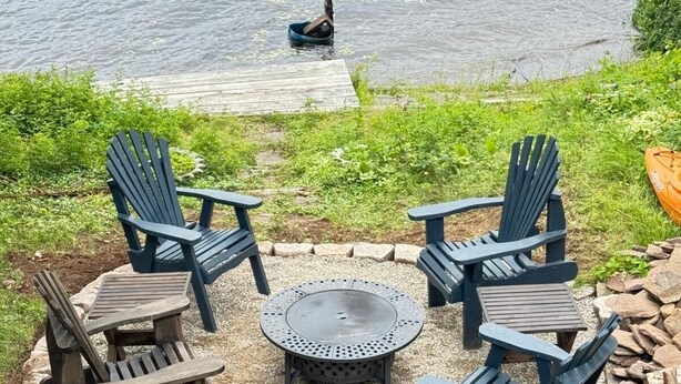 A circular outdoor seating area with Adirondack chairs and a fire pit, surrounded by a gravel border and a wooden deck extending into a lake. - Water front Getaway (Sault Ste. Marie)