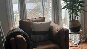A brown leather armchair with a white pillow and a throw blanket, positioned in front of a large window with white curtains. A potted plant is on a small round table beside the chair. - Water front Getaway (Sault Ste. Marie)