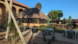 An outdoor seating area with tables and chairs, a thatched-roof structure, and palm trees.