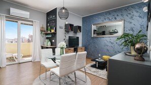 A modern living room with a glass dining table, white chairs, a TV, a coffee bar sign, and a large mirror.
