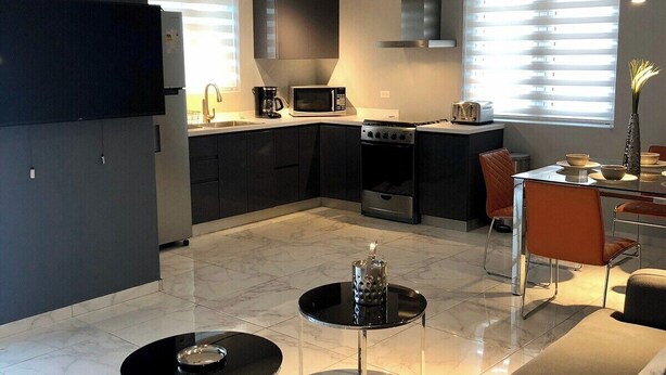 Een moderne keuken met een magnetron, fornuis en gootsteen. Er is een eethoek met een tafel en stoelen. De kamer heeft een groot raam met zonwering.