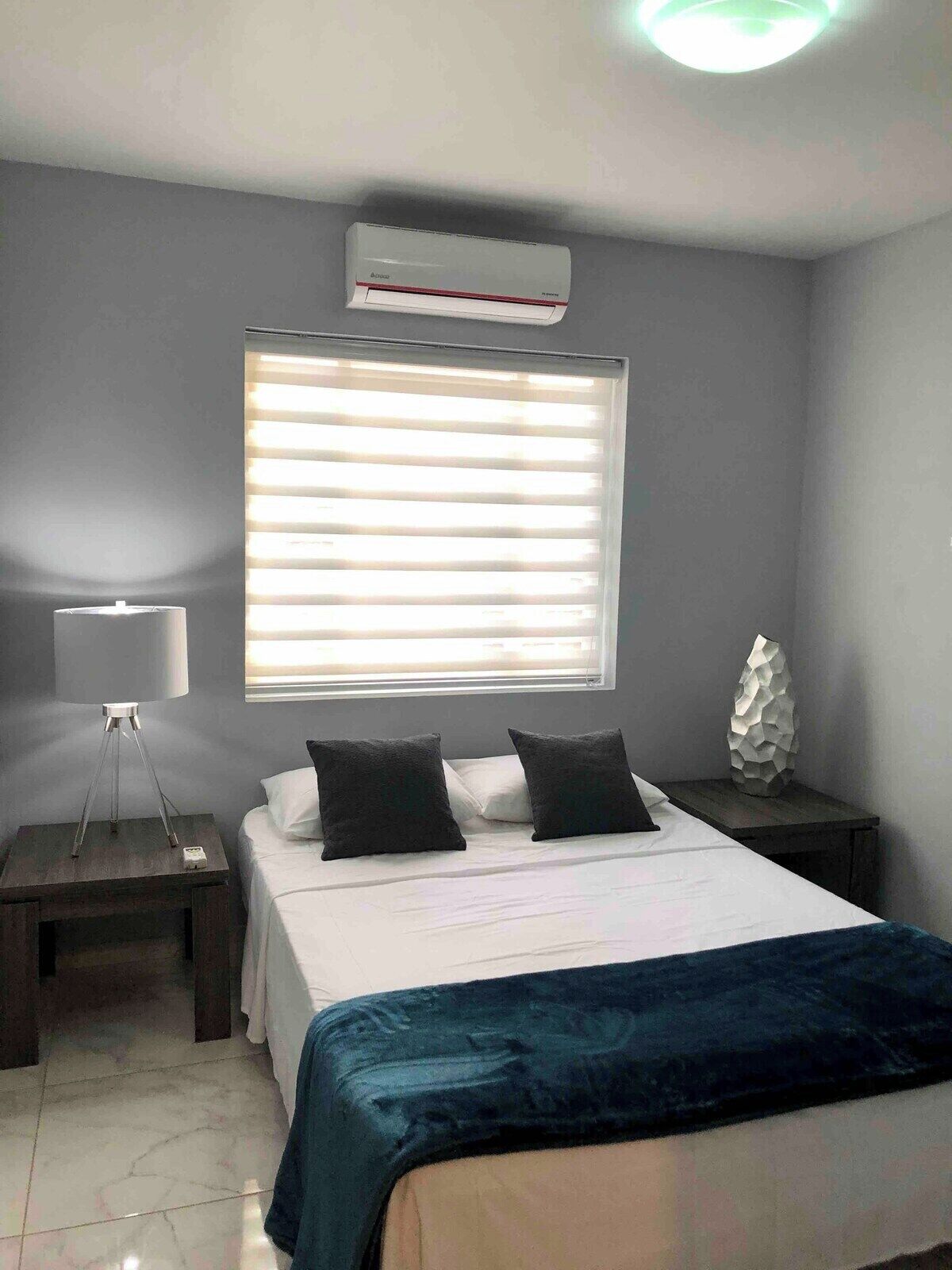 Une chambre à coucher comprenant un lit, une lampe de chevet, un climatiseur fixé au mur et une fenêtre avec des stores.