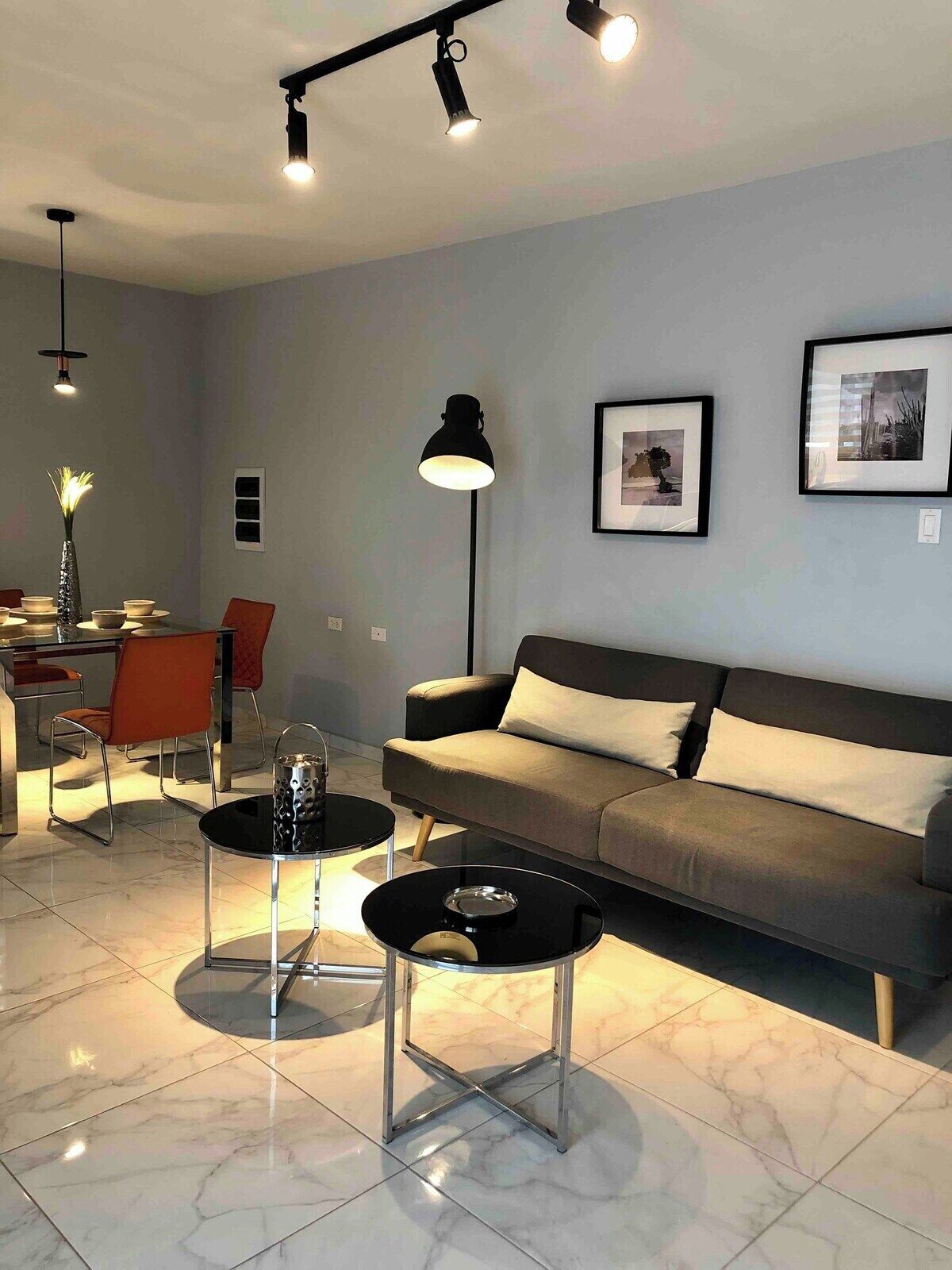 Een moderne woonkamer met een bank, twee bijzettafels, een eethoek met stoelen en een vloerlamp.