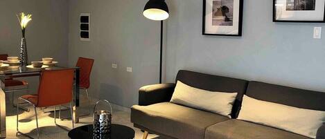 Een moderne woonkamer met een bank, twee bijzettafels, een eethoek met stoelen en een vloerlamp.