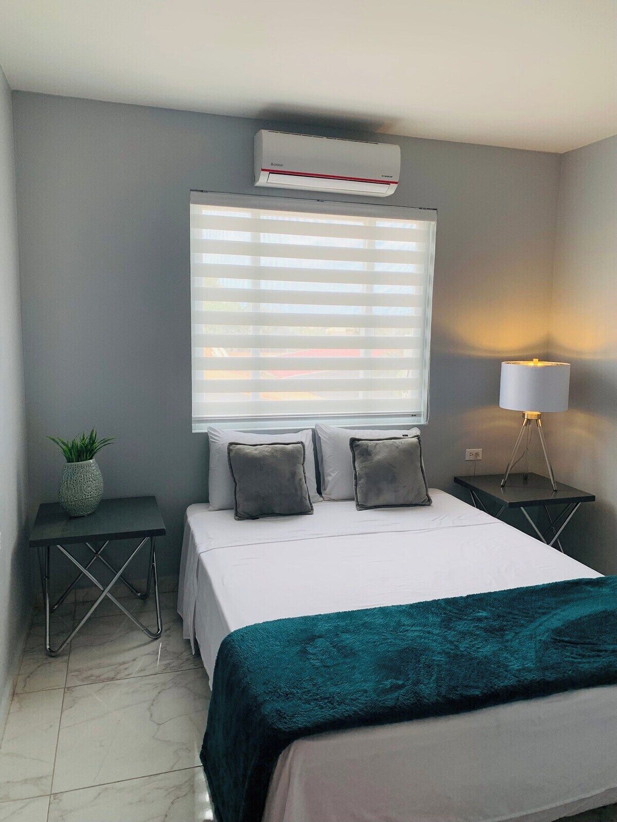 A bedroom with a bed, a window with blinds, a bedside lamp, a small table with a plant, and an air conditioning unit.