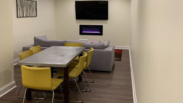 Een moderne woonkamer met een eethoek, een grijze bank, gele stoelen, een flatscreen-tv en inbouwspots.