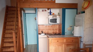 Cocina compacta con microondas, cafetera y un refrigerador pequeño. Se observa un armario de madera y un lavamanos. Una escalera es visible a la izquierda.