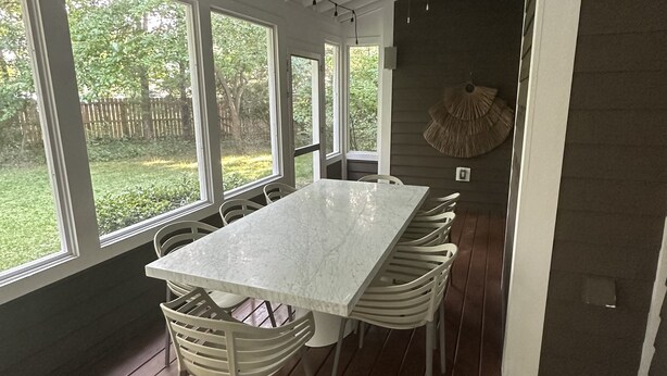 A covered patio with a marble table and white chairs, large windows, and a hanging clock.