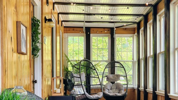 A room with wooden walls, a round table with a white top, wicker chairs, two hanging chairs with cushions, and large windows with greenery outside. - Riverside Historic Home near Downtown, Wineries, Hiking (Taylors Falls)
