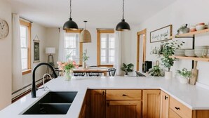 A modern kitchen with a central island, pendant lighting, wooden cabinets, and a dining area with a table and chairs. - Riverside Historic Home near Downtown, Wineries, Hiking (Taylors Falls)