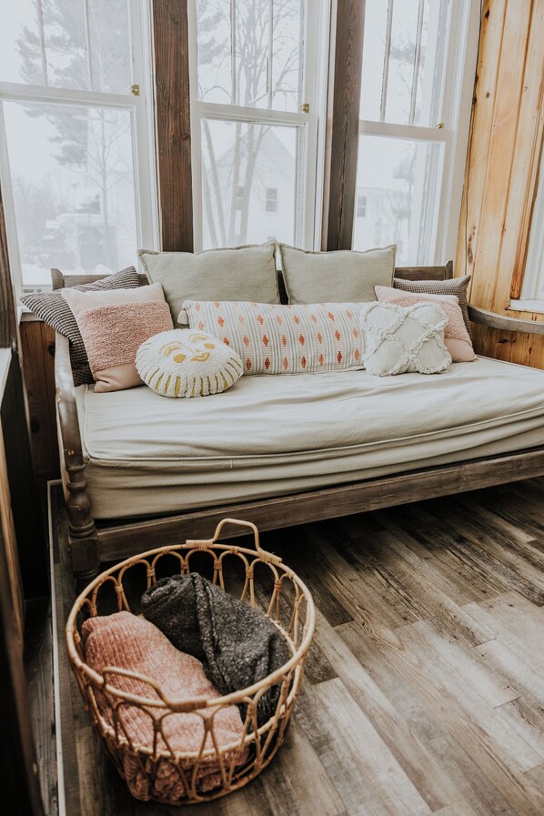 A cozy bedroom with a wooden bed frame, a wicker basket with blankets, and large windows with a view of trees.