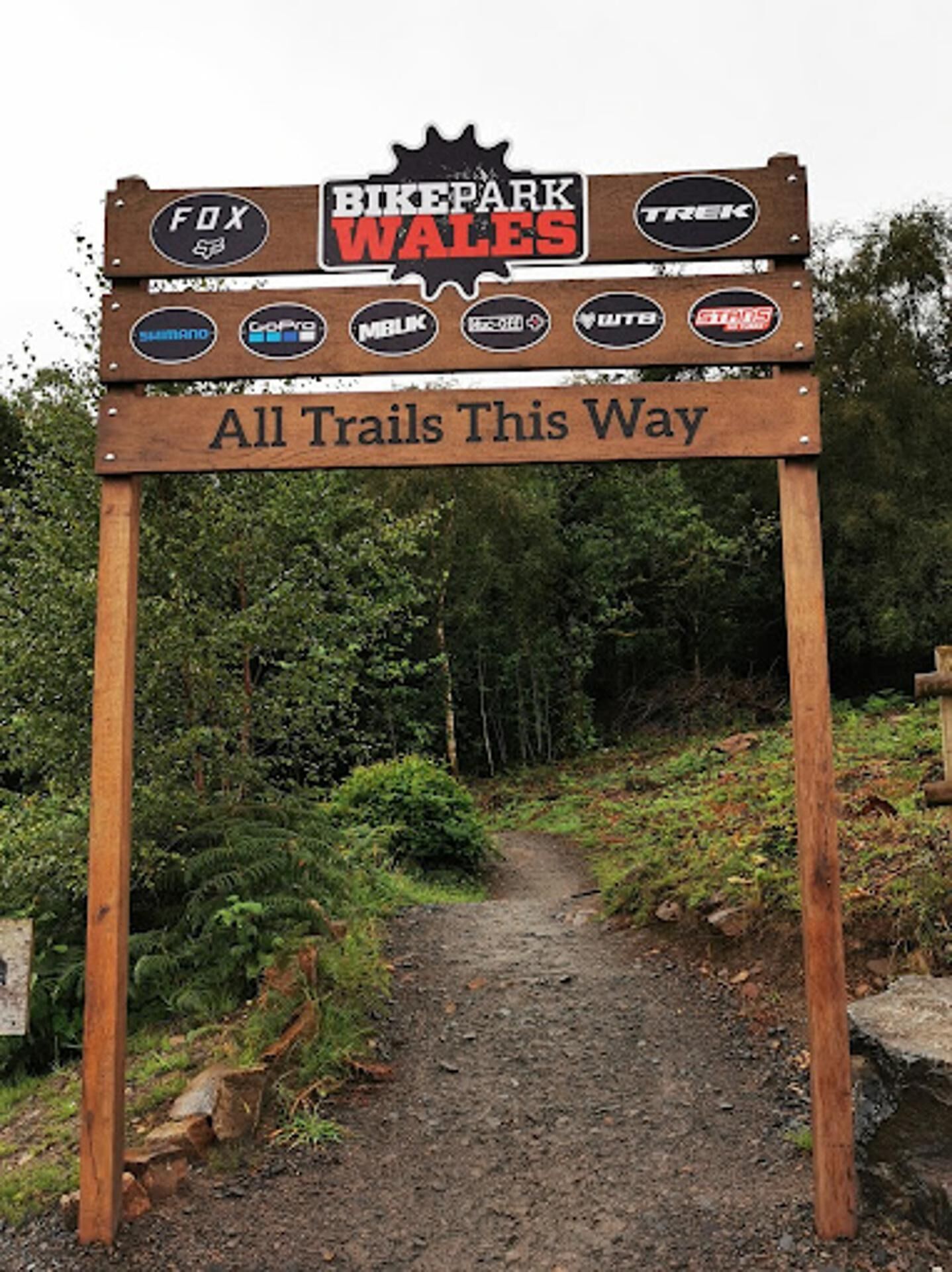 A wooden sign with multiple logos and the text "BIKE PARK WALES" and "All Trails This Way".