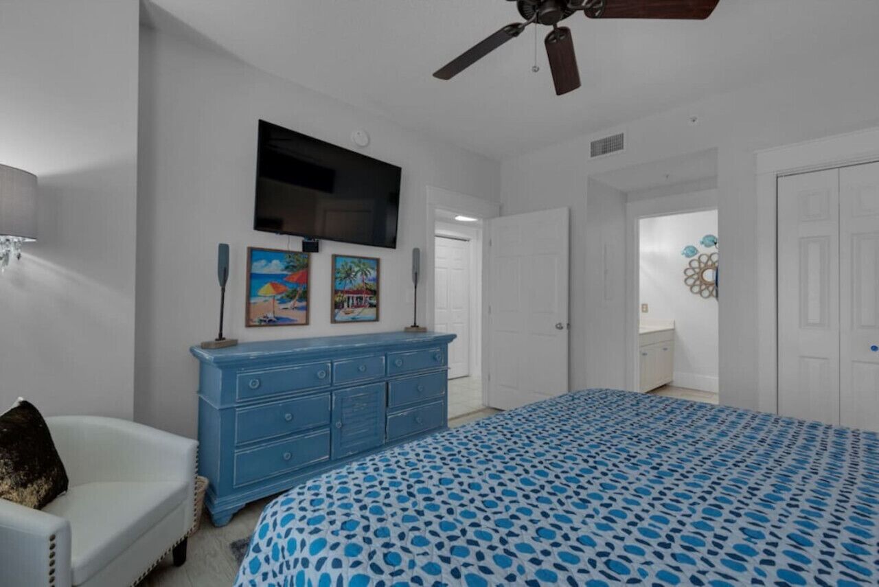 A bedroom with a bed, a dresser, a ceiling fan, and a TV mounted on the wall.