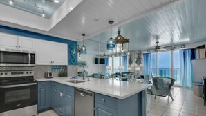 A modern kitchen with white cabinets, a stainless steel oven, and a dishwasher. There is a large island with a sink and a chair. The room has a blue and white color scheme and large windows with blue curtains. - Seaglitz - 2 bed 2 bath Oceanfront (Miramar Beach)