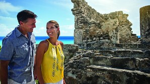 Ein Mann und eine Frau stehen in der Nähe alter Ruinen, im Hintergrund ein klarer, blauer Himmel und das Meer.