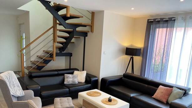 A modern living room with a black leather sofa, grey armchairs, a white coffee table, and a black floor lamp. A wooden staircase with a glass railing is visible in the background. - Maison Spacieuse Avec Piscine , Idéale Pour les Familles (Hilsenheim)