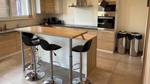 A modern kitchen with a central island, bar stools, stainless steel appliances, and a window with a towel hanging. - Maison Spacieuse Avec Piscine , Idéale Pour les Familles (Hilsenheim)