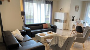 A modern living room with a black sofa, a grey armchair, and a white coffee table. There is a dining area with a white table and chairs in the background. - Maison Spacieuse Avec Piscine , Idéale Pour les Familles (Hilsenheim)