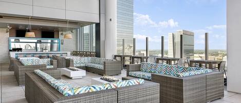 A rooftop lounge with wicker furniture, a bar area, and a city view.
