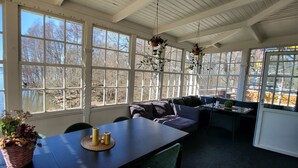 A sunroom with large windows overlooking a lake, a dining table with chairs, and a seating area with cushions. - Beautiful lake view house  (Vasteras)