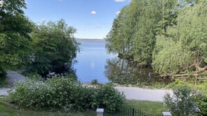 A view of a lake surrounded by dense trees and a paved path leading to a gated entrance. - Beautiful lake view house  (Vasteras)