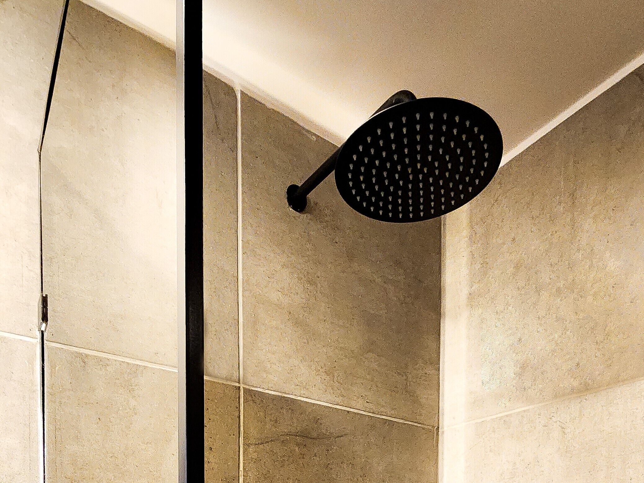 Un pommeau de douche doté d’une poignée noire et d’une tête circulaire, installé dans une salle de bain carrelée aux carreaux de couleur beige.