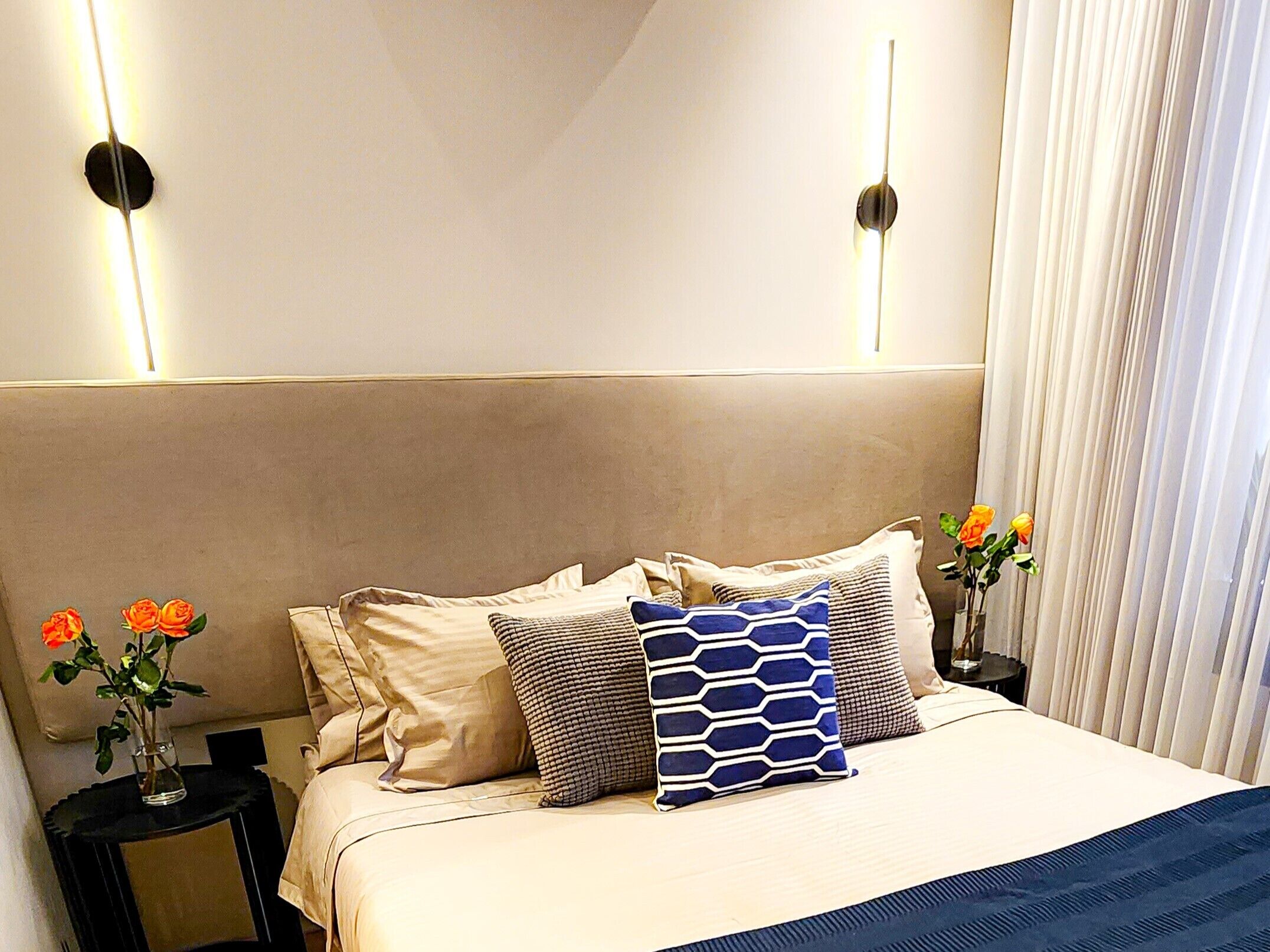 Un letto rifatto con cura, con una testiera beige, diversi cuscini e un cuscino azzurro decorato. Sono presenti due comodini con vasi di fiori.
