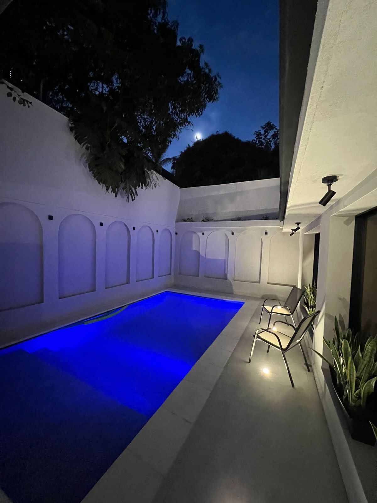 Una piscina con iluminación azul, rodeada de paredes blancas y un árbol.