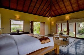 A bedroom with a large bed, wooden furniture, and large windows with open doors leading to a garden.