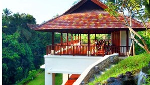 Uma casa com telhado de telhas vermelhas, varanda coberta e escadaria de pedra que leva a um jardim com lago e cachoeira.