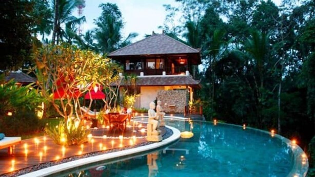 Uma piscina com borda curva, rodeada por plantas tropicais e um pequeno edifício com telhado em azulejos.