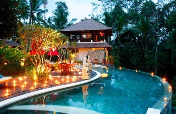 Relax by the serene pool surrounded by lush tropical vegetation and a traditional-style building.