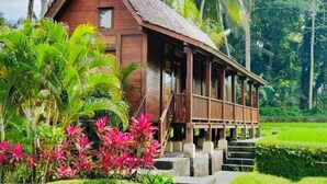 Uma casa de madeira com telhado de palha, cercada por vegetação exuberante e palmeiras.