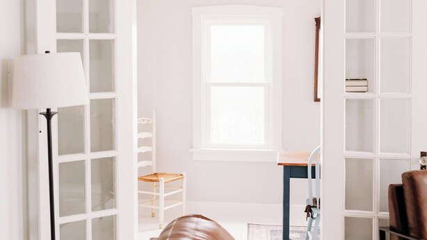 A modern living room with a brown leather sofa, a lamp, a bookshelf, and a window.