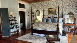 A bedroom with a four-poster bed, a wooden desk, a chair, and a wicker chair with a magazine.