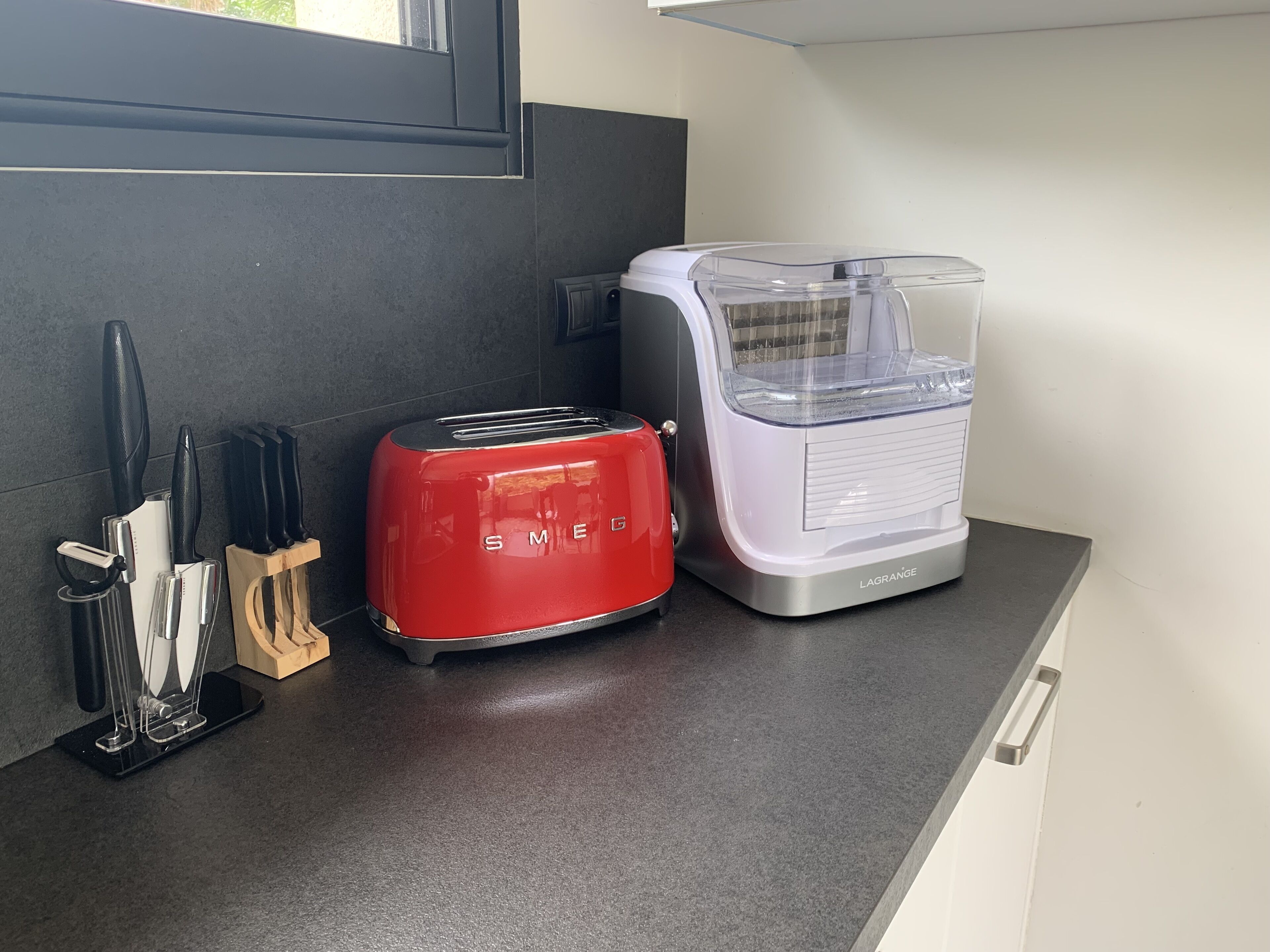 A kitchen counter with a red toaster and a white appliance, possibly a food processor, with a knife block and utensils to the left.