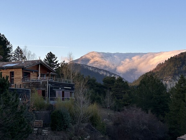 A wooden cabin with a balcony, surrounded by dense forest and mountains.