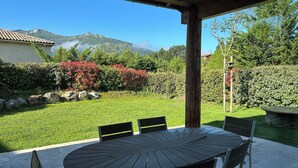 Un patio exterior cubierto con una mesa redonda y sillas, rodeado de un césped y jardín bien cuidados con diversas plantas y setos.