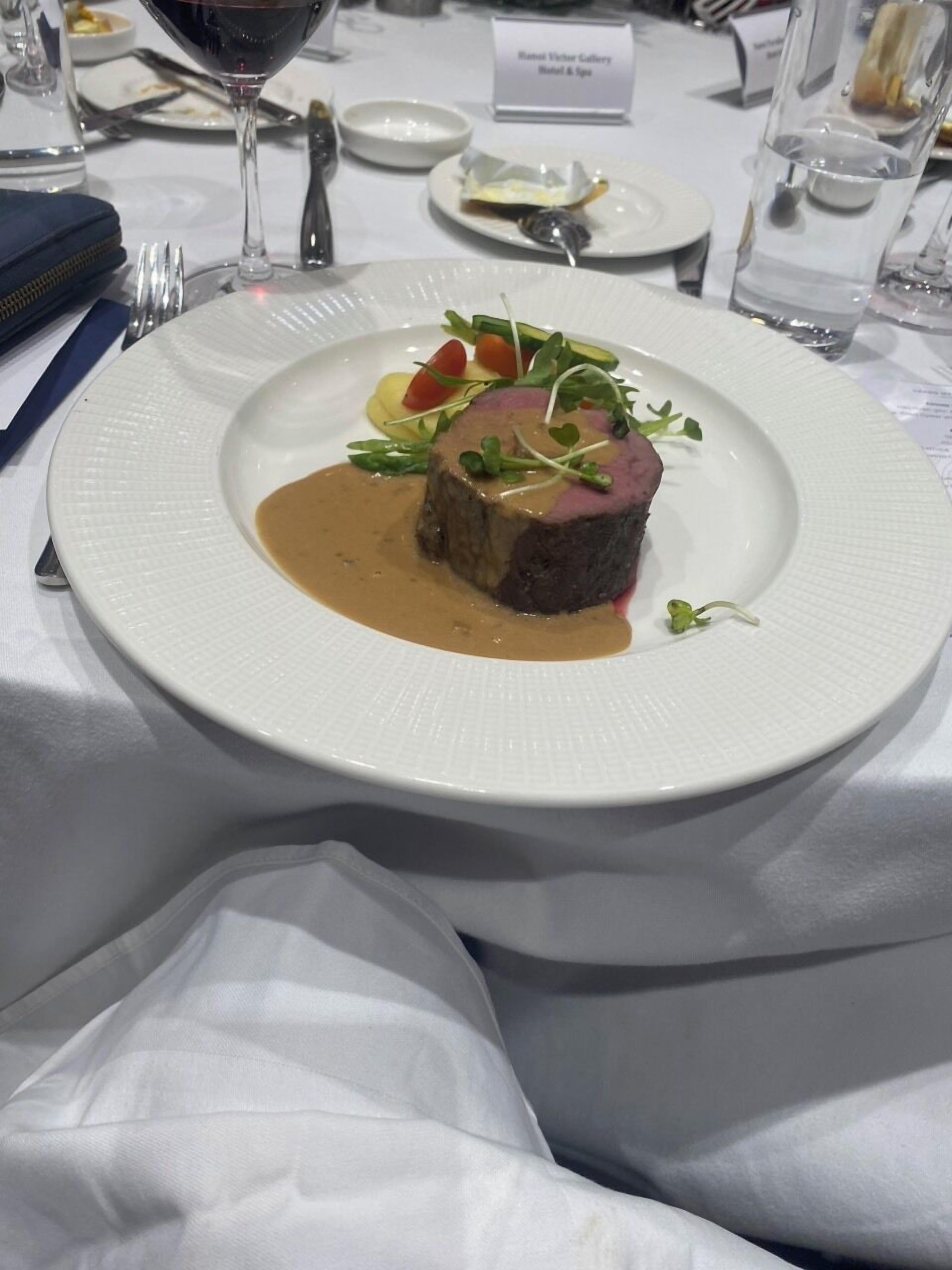 A white plate with a steak topped with greens and a brown sauce, set on a table with a white tablecloth.