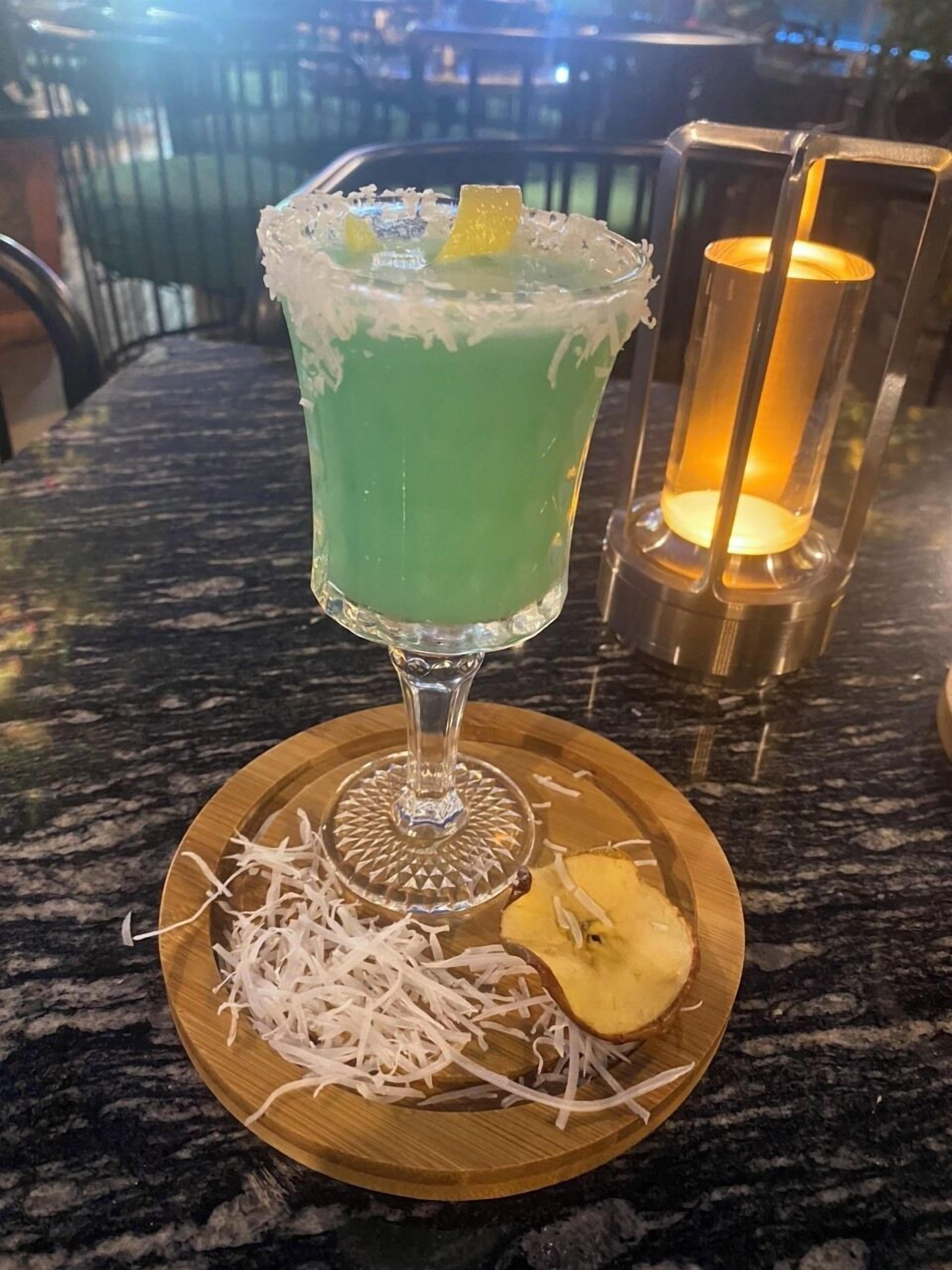 A cocktail in a glass with a lime garnish, served on a wooden coaster with shredded white material and a pineapple slice.