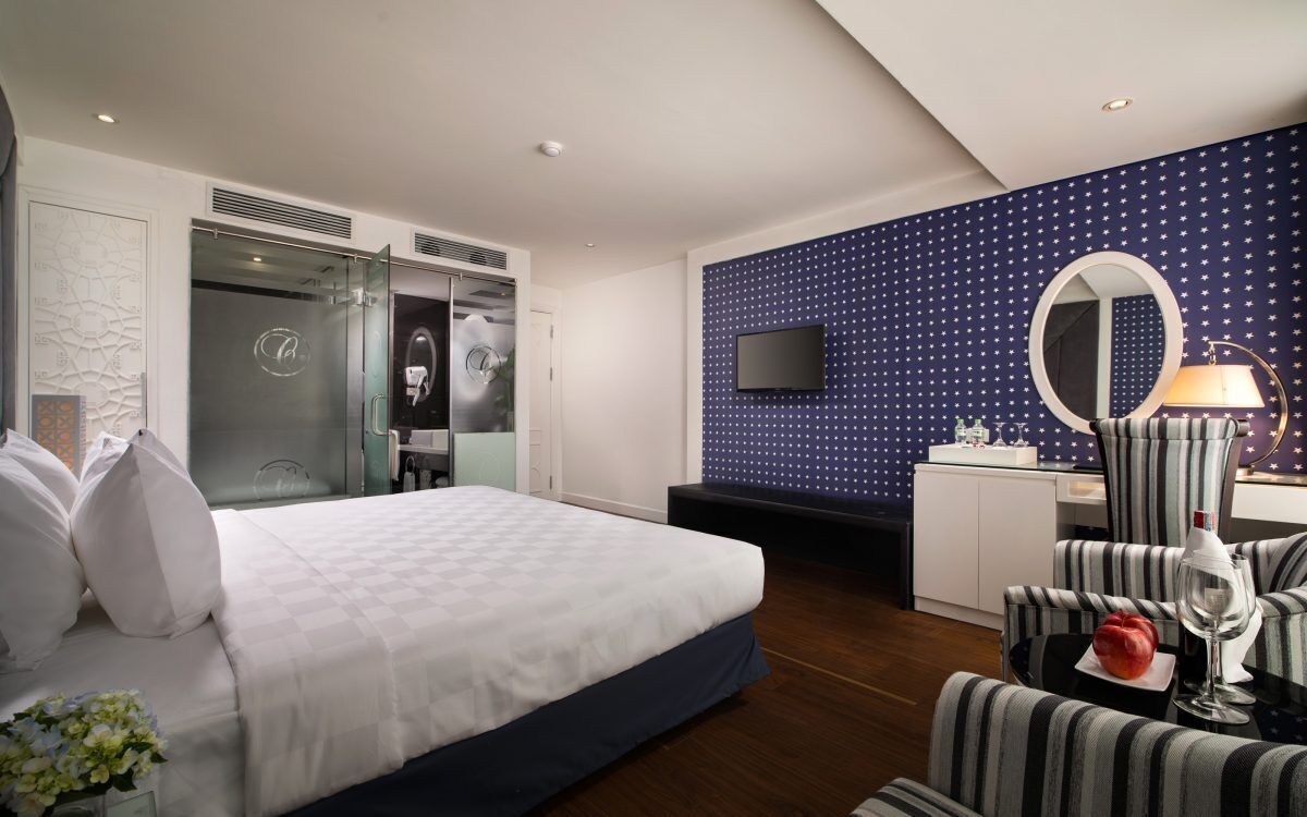 A modern hotel room with a large bed, a desk with a computer, a TV, and a seating area with a striped chair and a lamp.