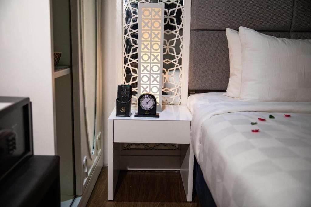 A neatly made bed with white bedding and a decorative headboard, a white bedside table with a clock and a remote control, and a patterned wall divider.