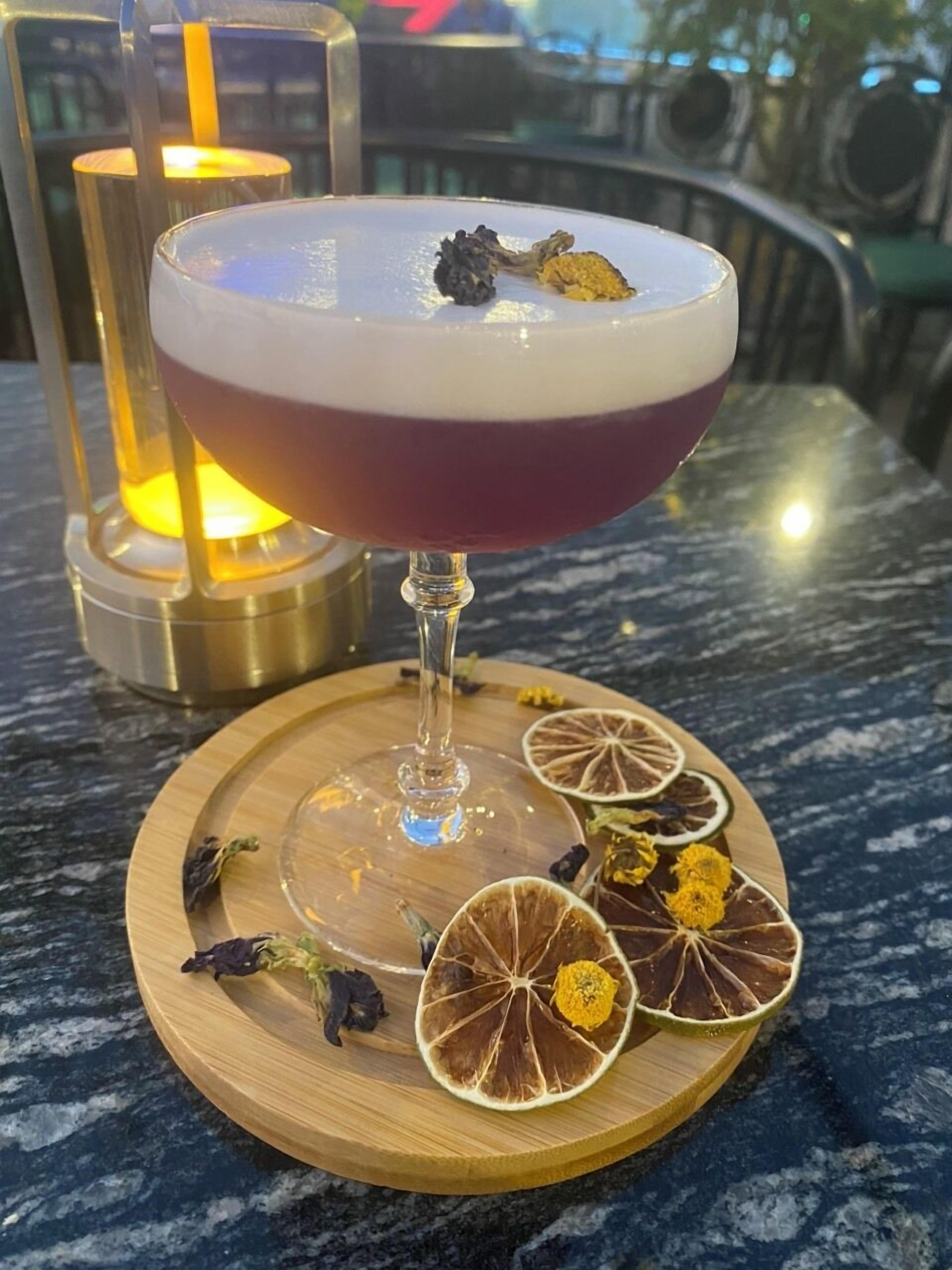 Un cocktail servi sur une planche en bois, accompagné de tranches d’agrumes séchées et de fleurs, d’une bougie et d’un verre.