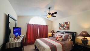 Ein Schlafzimmer mit Bett, Deckenventilator, Fernseher, Lampe und einem Fenster mit Vorhängen.